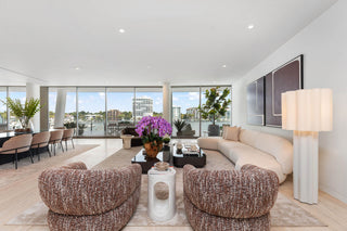 Home Staging Miami - A modern living room interior with a curved cream sofa, two textured brown armchairs, a white side table, a floor lamp, and large windows overlooking a waterfront view.