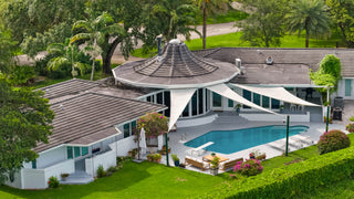 Home Staging and Turnkey Furniture Rental - Aerial view of a modern luxury estate featuring a distinctive flying-saucer style pavilion with a tiled dome roof, white curved walls, a swimming pool, and expansive white canopy screens over a lush green lawn.