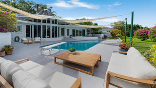 Home Staging and Turnkey Furniture Rental - Outdoor patio area with a swimming pool, modern white house in the background, and light wood lounge furniture with white cushions under a beige canopy.
