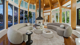 Home Staging and Turnkey Furniture Rental - A luxury modern living room featuring two curved, low-profile modular sofas in light beige and gray fabric, arranged around a round white coffee table on a large beige area rug. The room has a high arched wooden ceiling, wood floors, and large windows looking out to a pool.