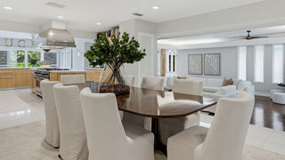 Home Staging and Turnkey Furniture Rental - A modern dining room featuring a dark wood oval table surrounded by white upholstered chairs, with a large green leaf arrangement in the center. The room is connected to a bright kitchen and living area with light gray walls, recessed lighting, and large windows.