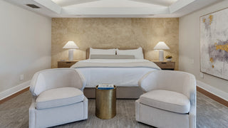 Home Staging and Turnkey Furniture Rental - A modern bedroom interior featuring a neatly made bed with white linens, two upholstered armchairs facing a small round gold side table, and a textured beige accent wall behind the bed.