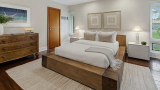 Home Staging and Turnkey Furniture Rental - A modern bedroom featuring a low wooden platform bed with white bedding, matching white nightstands, and a light beige rug on dark wood floors. The room has light gray walls, a wooden dresser on the left, and two windows on the right.